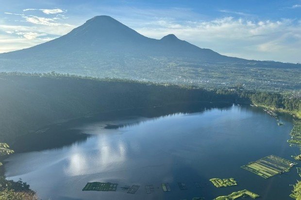 Pemandangan Telaga Menjer Wonosobo dengan latar belakang gunung yang biru dan langit cerah, serta pantulan awan di permukaan air dan keramba ikan di pinggir danau.