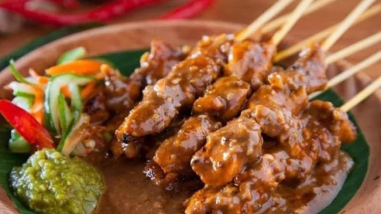 Sate Madura dengan bumbu kacang kental disajikan di atas piring, terlihat beberapa tusuk sate dengan potongan daging yang dibakar, dilengkapi sambal dan lalapan.